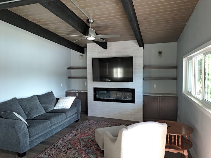 Sebule ya ghorofa iliyo na televisheni mahiri ya "60", meko ya umeme, feni ya dari na kochi kubwa na kiti cha kukaa. Chumba hiki kinaelekea kwenye sitaha yenye mwonekano wa mto hapa chini