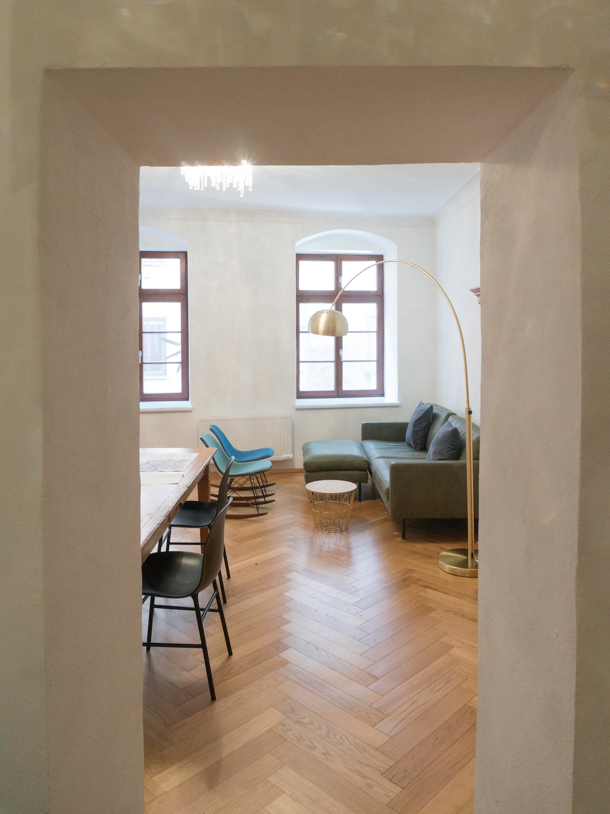 A spacious living area is visible through an archway. The room features hardwood flooring arranged in a herringbone pattern and is furnished with a green sofa, a light-colored side table, and a mix of modern chairs. Two windows allow natural light to illuminate the space.