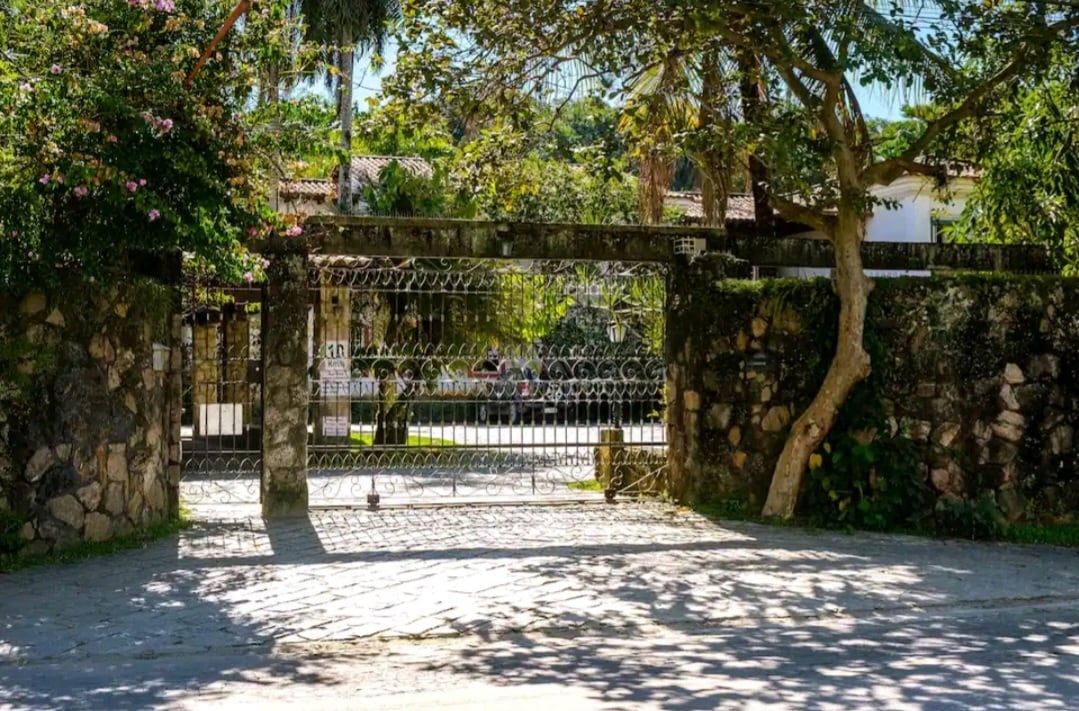 A decorative stone gate stands amid lush greenery, framed by trees and flowering plants. Cobblestone ground leads up to the entrance, where a hint of sunlight filters through the foliage, creating layered shadows on the path.
