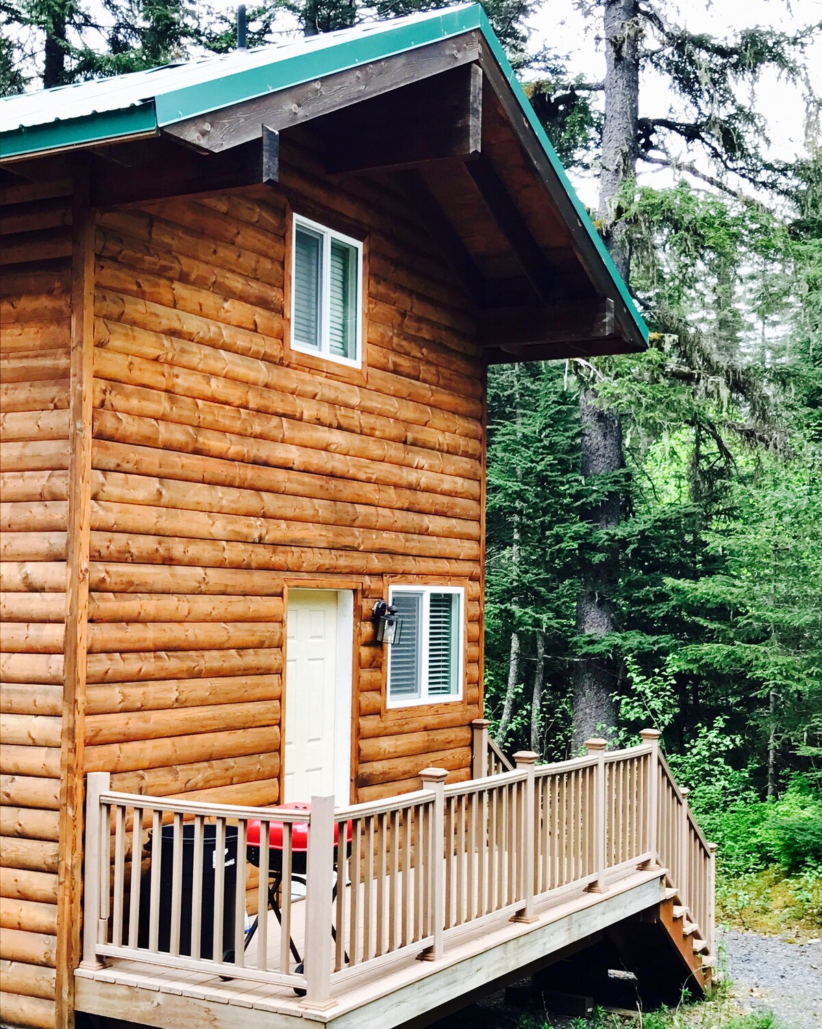 A wooden cabin with a sloped roof is surrounded by lush greenery. The entrance features a white door and a small porch with a wooden railing. A red table can be seen on the porch, inviting guests to enjoy the outdoor space.