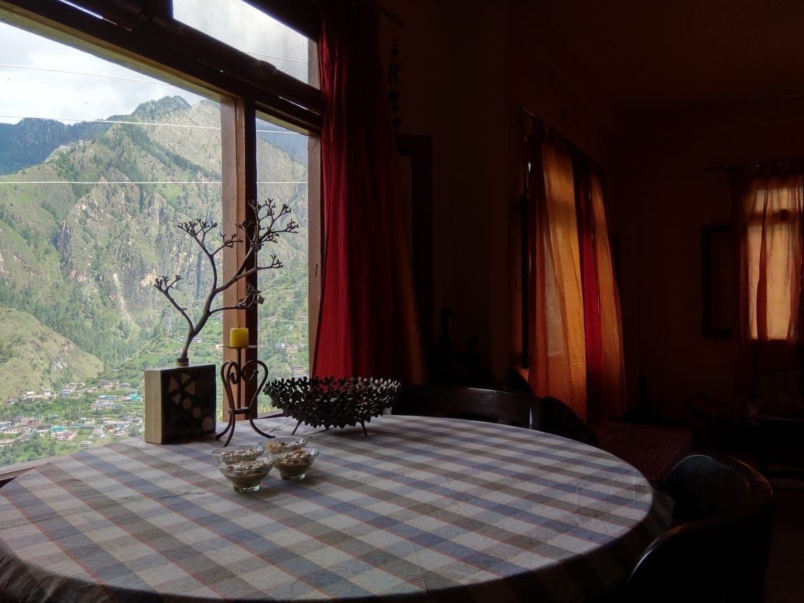 A round table is set against a backdrop of large windows, revealing mountain views. Decorated with a vase and candles, the table complements the space, while vibrant curtains frame the windows, allowing natural light to illuminate the room.