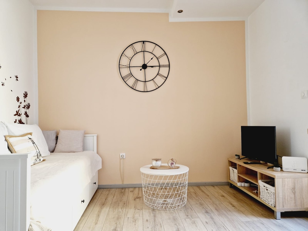A neutral-toned living area features a wall clock as a centerpiece above a light-colored sofa. A low coffee table with a decorative item sits on a wooden floor, while a television is positioned on a side table, providing a functional yet simple layout.