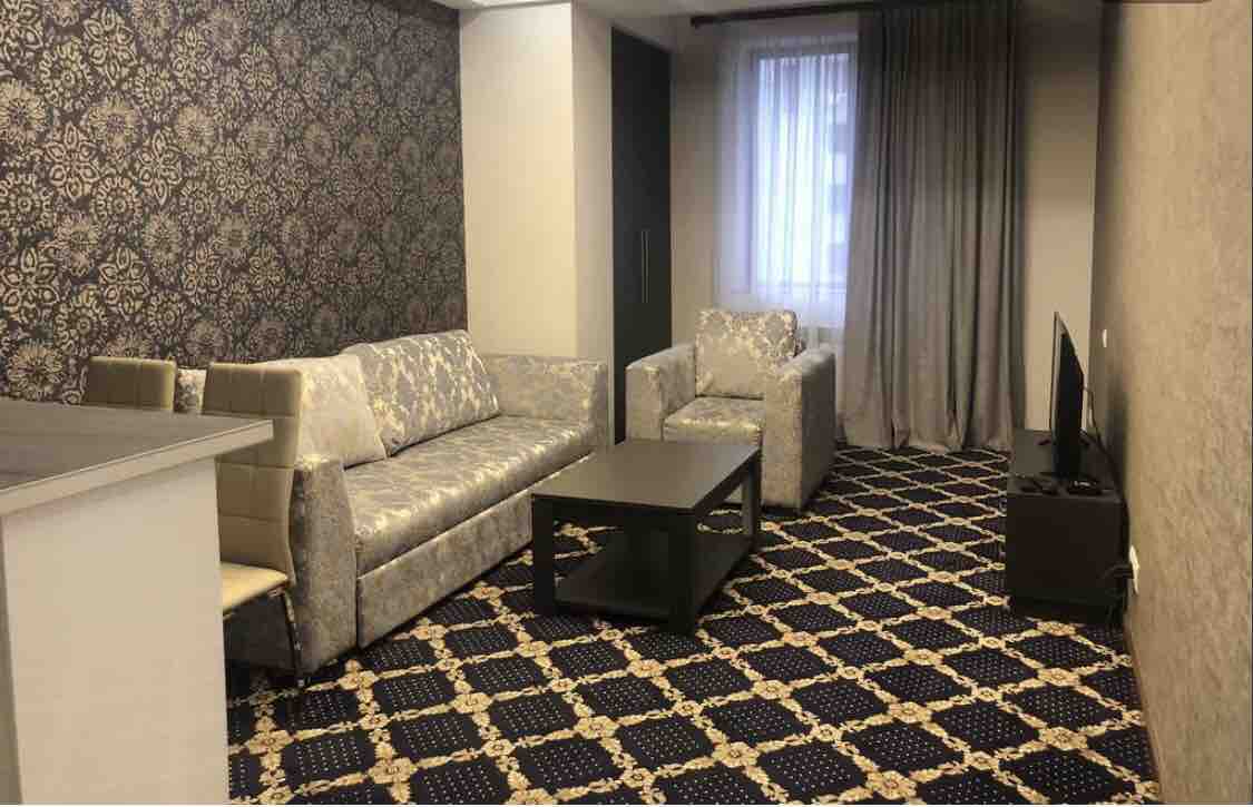 A stylish living area features a patterned carpet and textured wallpaper. Comfortable seating is provided by a spacious sofa and two armchairs. A dark coffee table sits in the center, while a television is positioned against the wall. Natural light filters in through a nearby window, dressed in soft curtains.
