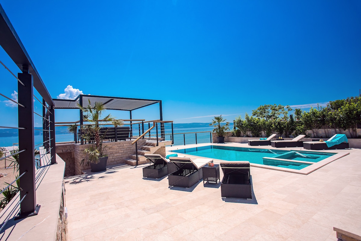 An outdoor sun deck features a private, heated pool with an infinity edge and a hydromassage area. Six lounge chairs are positioned for relaxation, with a shaded dining area and views of the sea visible in the background.