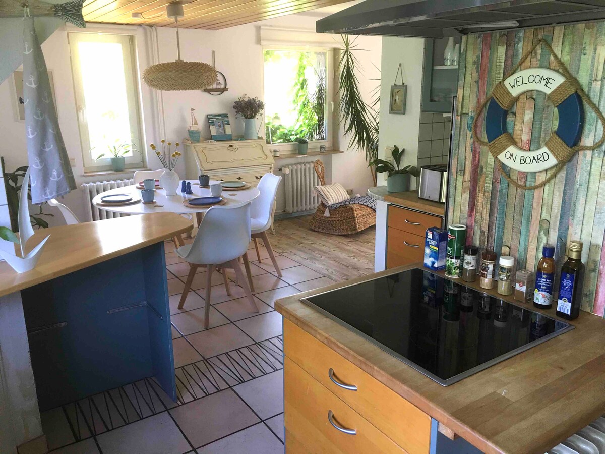 A modern kitchen area is featured, with an island counter, an induction cooktop, and a variety of cooking oils and spices displayed in a line. Light enters through multiple windows, illuminating the space that includes a dining table set for four and decorative coastal-themed accents.