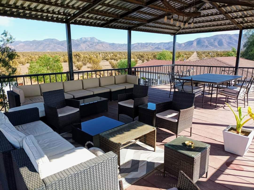 An expansive rooftop terrace features comfortable seating with a sectional sofa and multiple chairs surrounding a glass table. The area offers panoramic views of the surrounding mountains and landscape, under a covered roof that provides shade.