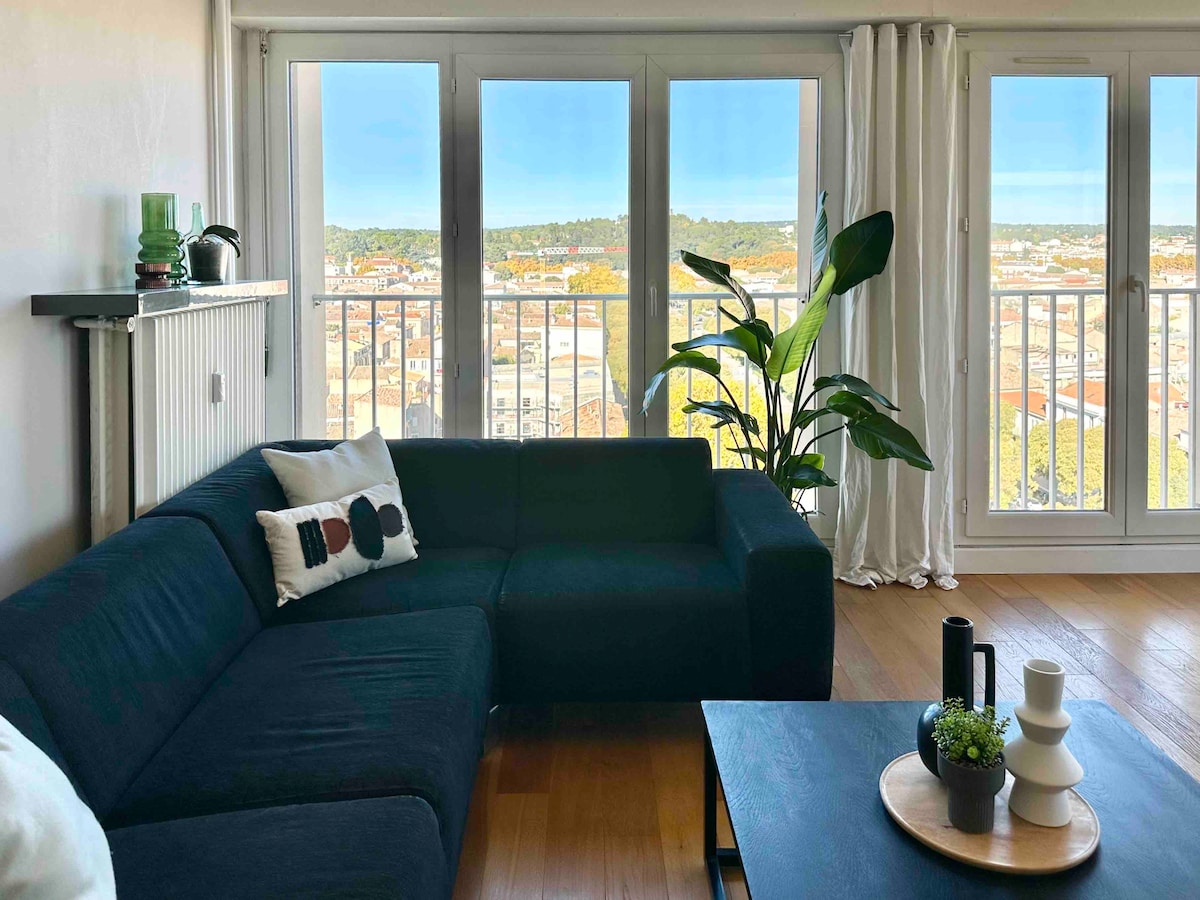 A spacious living area features a dark sectional sofa adorned with a decorative pillow, complemented by a light-colored coffee table. Large glass doors provide an expansive view of the cityscape, with greenery and natural light enhancing the atmosphere. A tall potted plant adds a touch of nature.