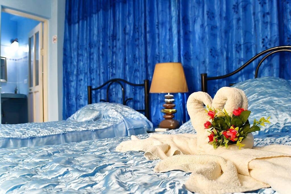 The room features two beds draped in soft, blue bedding, complemented by a decorative arrangement of towels shaped like a heart, accompanied by a small bouquet of flowers. A bedside lamp casts a gentle glow, and a bathroom area is visible in the background.