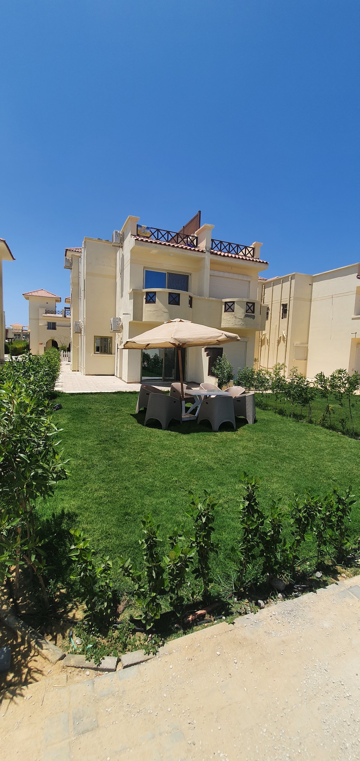 A spacious outdoor area is presented, featuring a private garden with well-maintained grass and hedges. A shaded seating area with a gazebo is visible, surrounded by lush greenery. The villa's yellow exterior provides a bright backdrop under a clear blue sky.