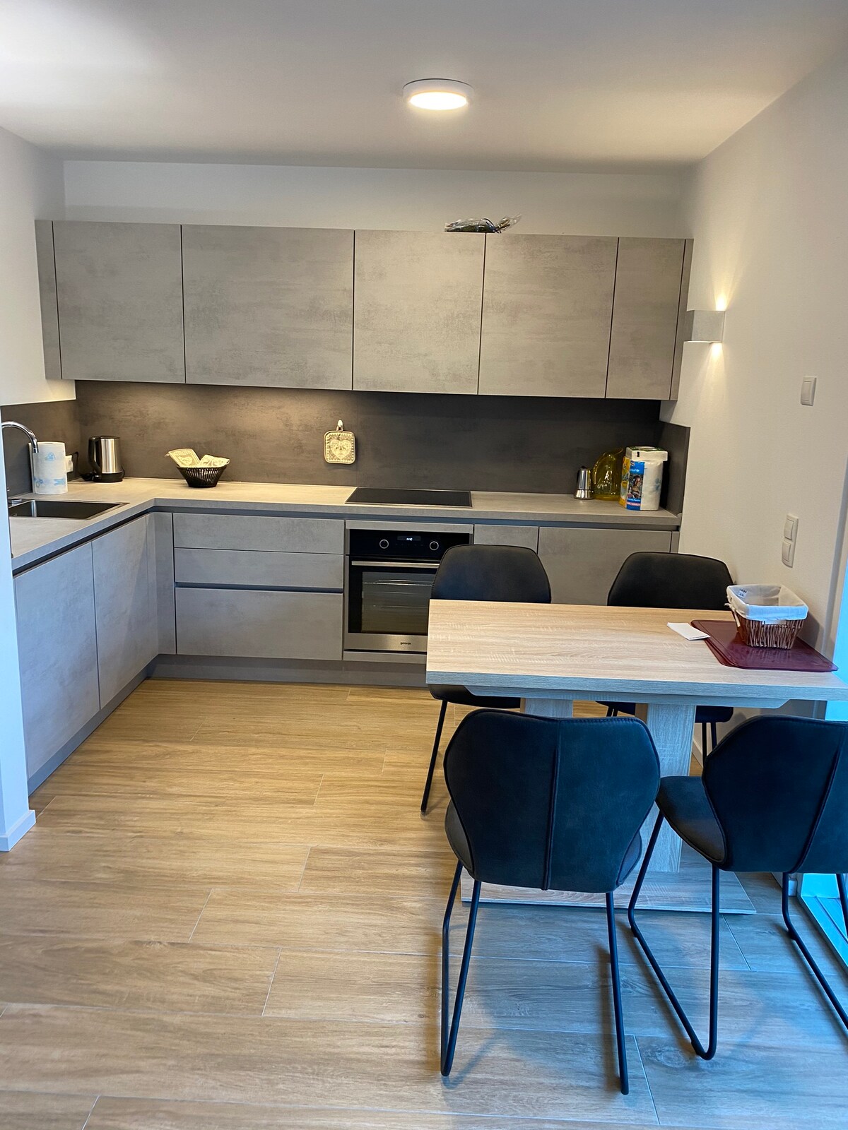 The kitchen features modern cabinetry with a clean design, complemented by a sleek countertop and built-in appliances. A wooden dining table is surrounded by four black chairs. Natural light enhances the ambiance from a window, contributing to the space's functionality.