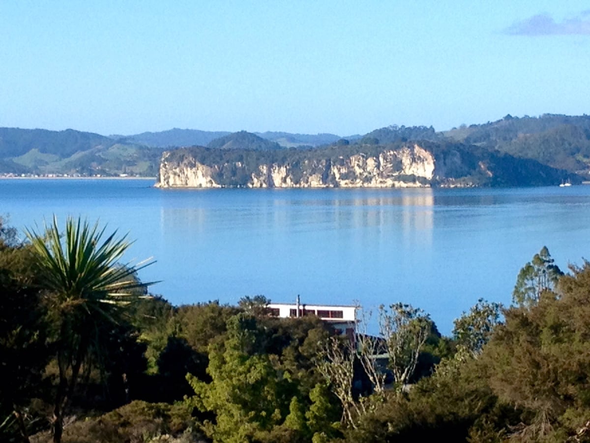A tranquil sea view is presented, featuring a coastal cliff rising above calm waters. Lush greenery frames the scene, while distant hills are visible in the background. The sky above is clear with soft blue hues, creating a serene atmosphere.