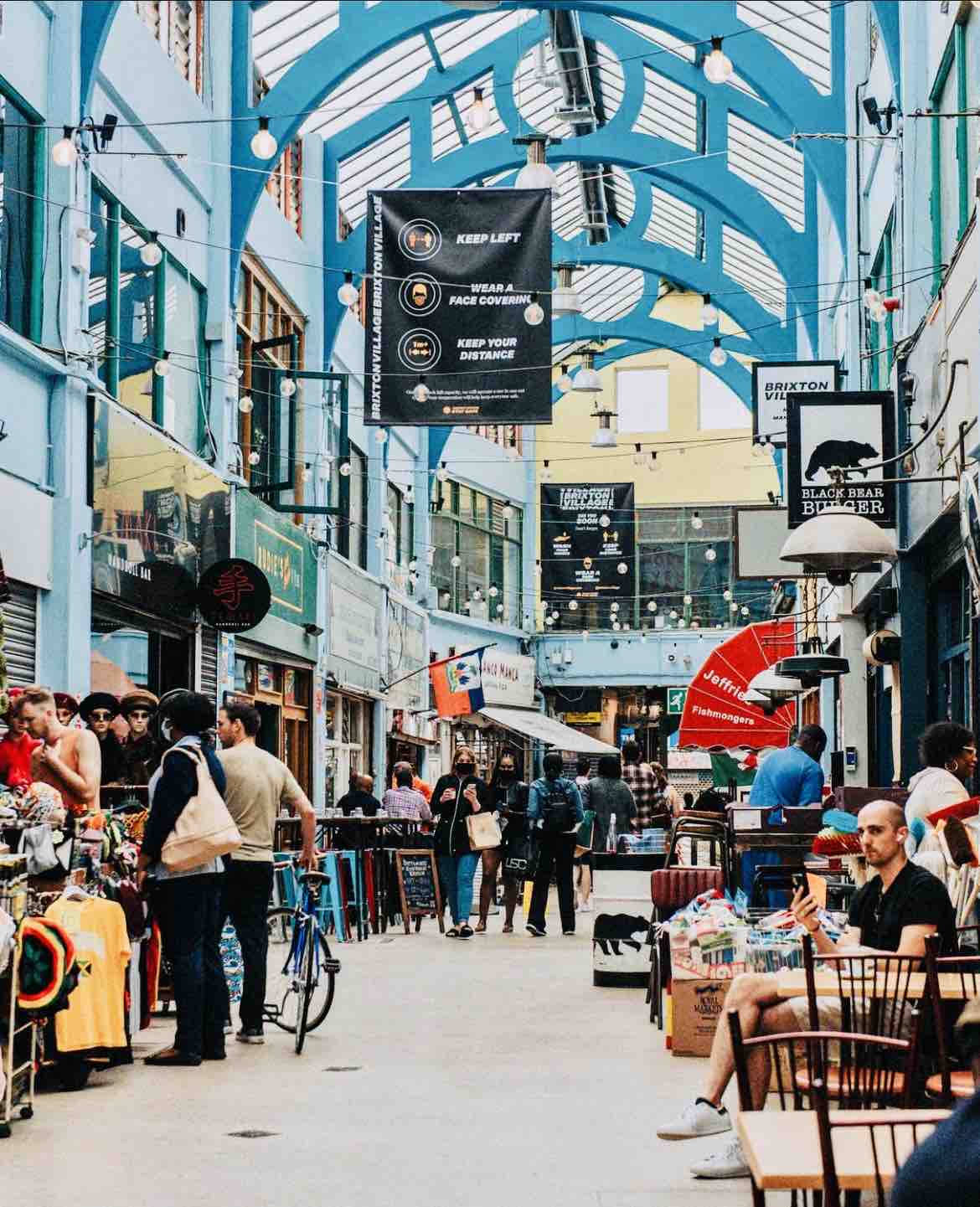 An indoor market area features high blue ceilings with natural light streaming through skylights. Stalls display a variety of items, while visitors browse the shops. Tables and chairs are positioned along the sides, creating a lively space for socializing. Decorative signage adds to the vibrant atmosphere.