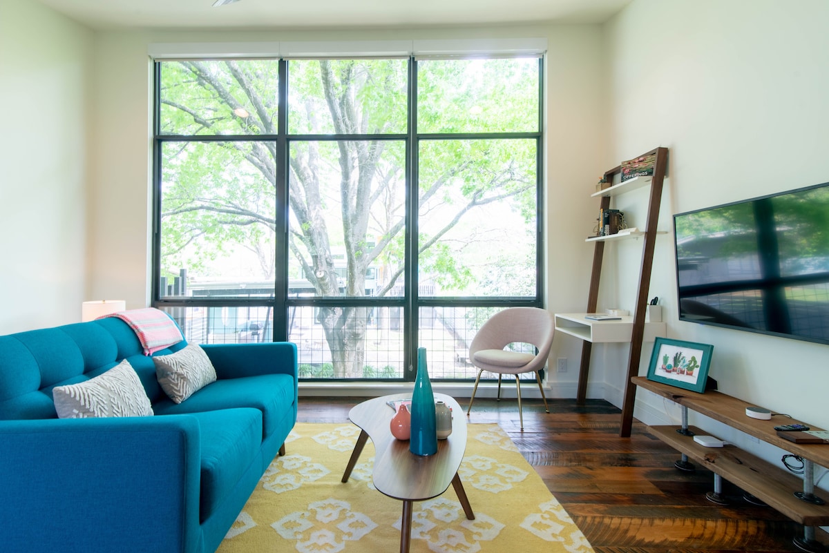A modern living area features a bright blue sofa adorned with decorative pillows, complemented by a low wooden coffee table. Large windows provide a view of lush green foliage outside. A minimalist shelf and TV stand enhance the contemporary design of the space.