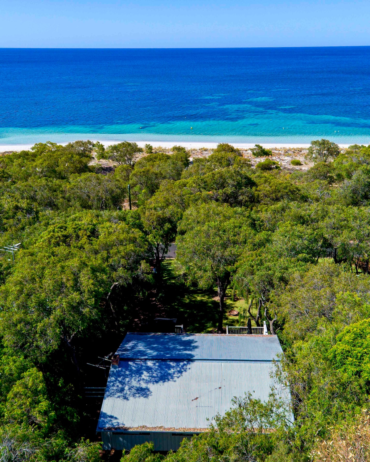 Aerial view showcasing the house nestled among lush greenery, with the vivid blue ocean and sandy beach in the background. The clear waters gently transition to sandy shores, highlighting the natural beauty of the Quindalup waterfront.