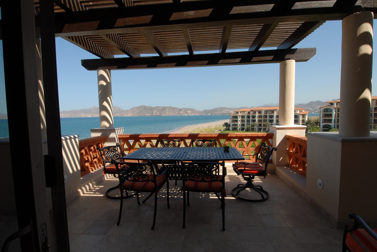 A spacious terrace offers a dining area with a large table and several chairs, providing stunning views of the coastline and the sea. The pergola structure allows natural light to filter in while providing some shade. Coastal hills are faintly visible in the background.
