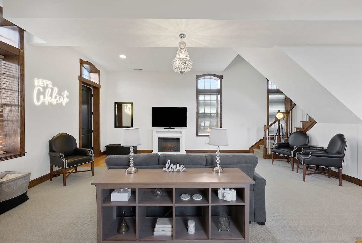 A spacious living area is highlighted by a contemporary chandelier and a cozy faux fireplace. Modern black chairs are positioned around a central grey table, which features decorative items. Large windows allow natural light to fill the room, enhancing its bright and inviting ambiance.
