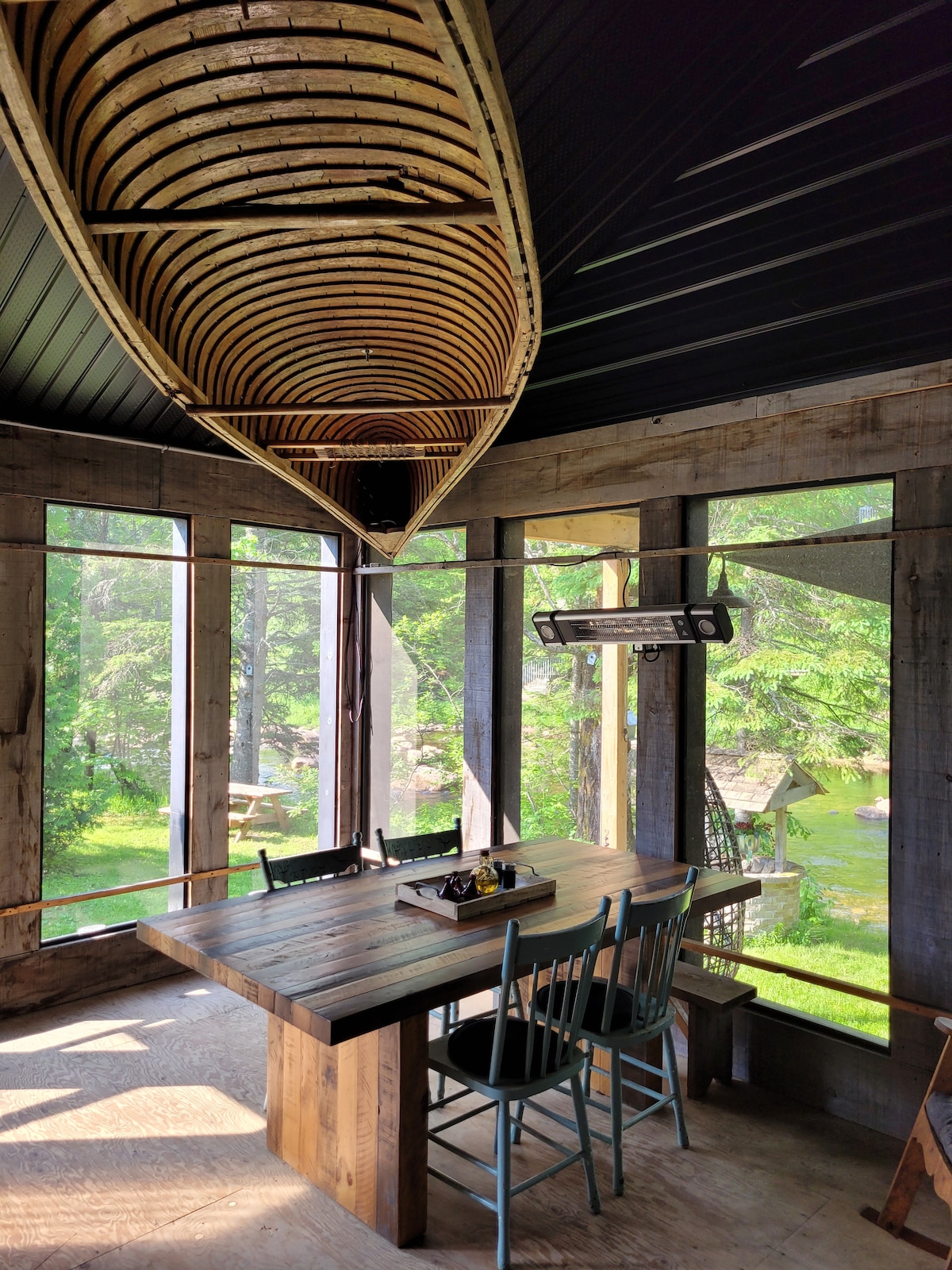 A dining area is featured under a wooden ceiling, with a large table surrounded by rustic chairs. A canoe hangs above, adding a unique touch. Sunlight filters through large windows, providing views of the lush outdoor scenery and enhancing the natural ambiance.
