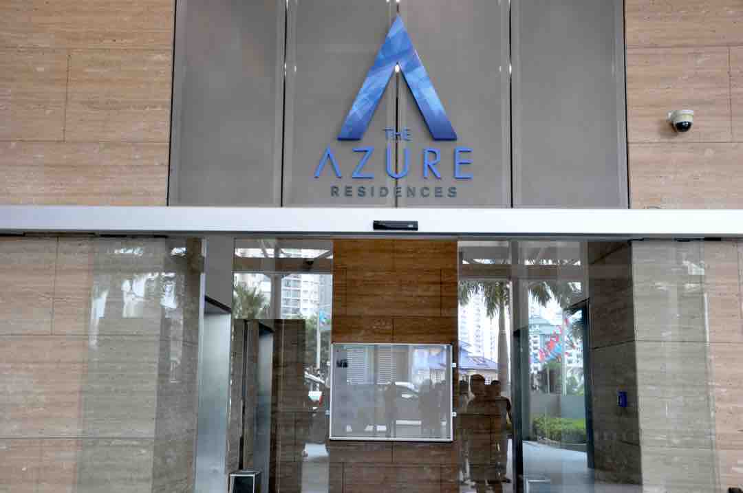 The entrance of The Azure Residences is shown, featuring a modern glass door with a prominent blue logo. Natural stone walls create an elegant backdrop, while indoor lighting enhances the welcoming ambiance. Palm trees are visible outside, suggesting a tropical environment.