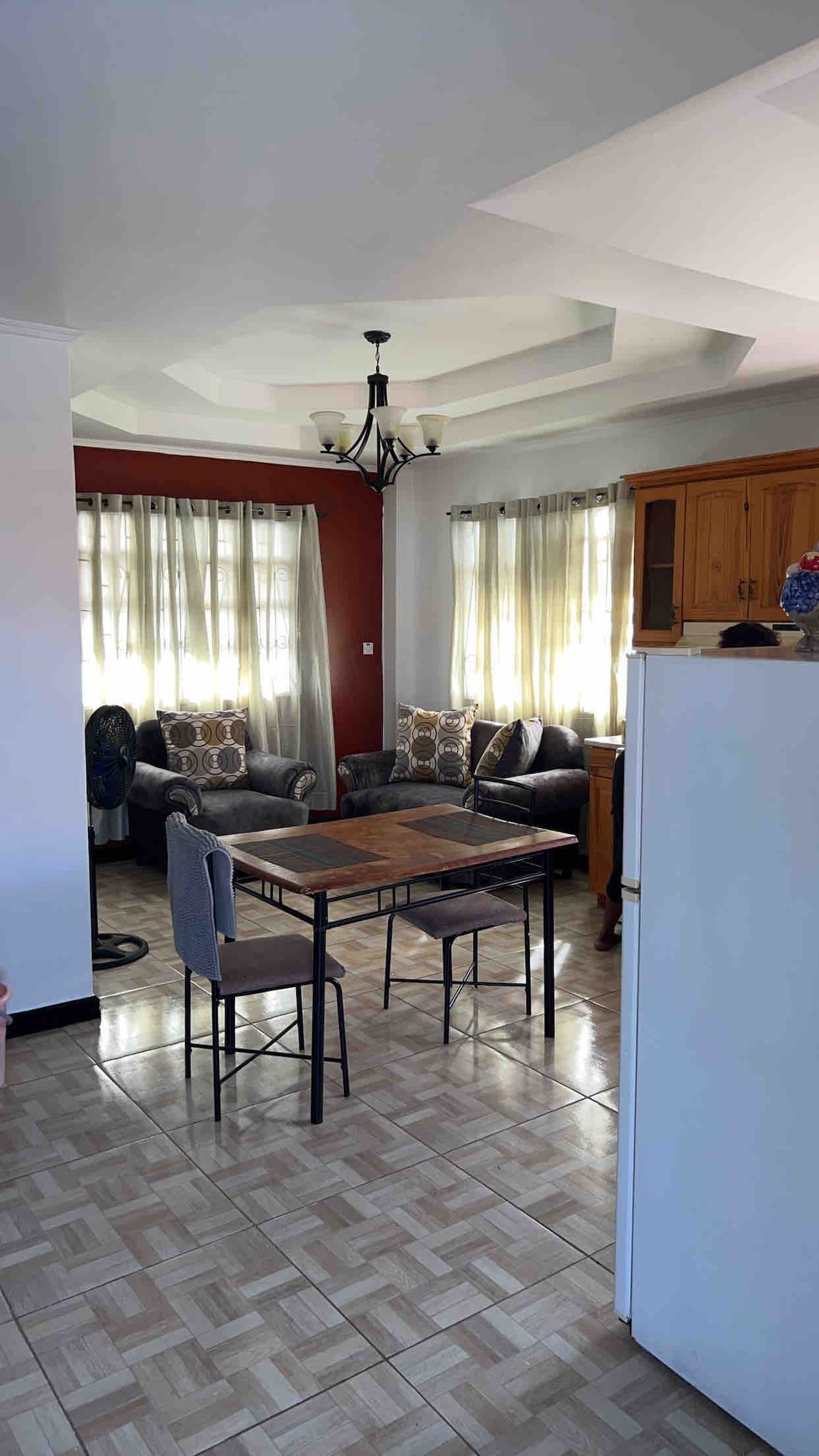 An inviting dining area features a wooden table surrounded by two chairs. Nearby, a cozy seating arrangement includes two gray upholstered chairs with accent pillows. Soft natural light filters through sheer curtains, highlighting the warm tones of the walls and the tiled floor.