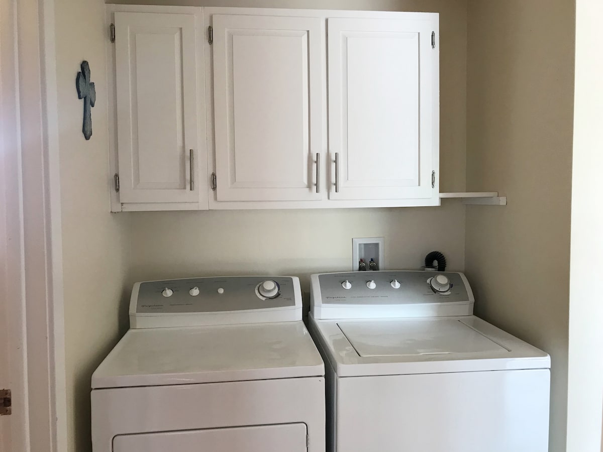 A practical laundry area features a pair of appliances, including a washing machine and dryer, placed side by side. Above, white cabinets provide storage space, while a shelf offers additional utility. The walls are painted in a soft color, enhancing the room's functionality.