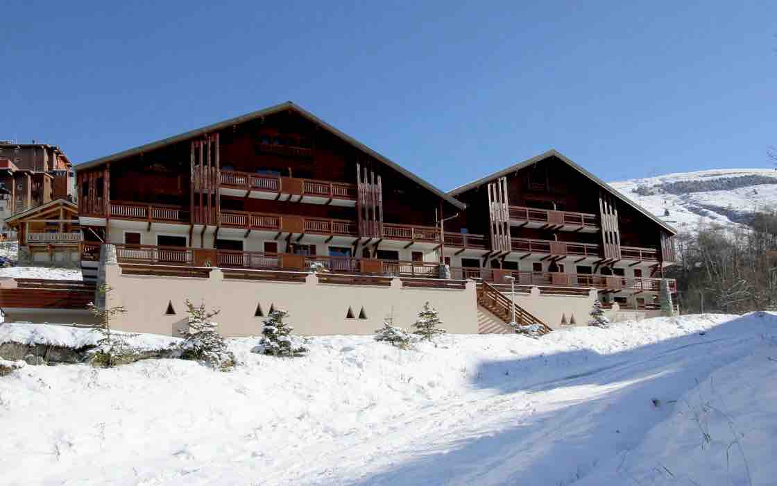 A two-building chalet-style residence is framed by snow-covered ground. Each building features multiple balconies with wooden railings and a sloping roof. Surrounding greenery adds contrast against the winter landscape, highlighting the remote mountain setting.