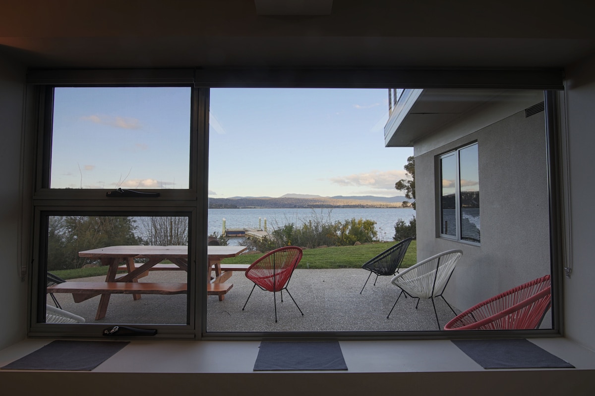 A large window frames a scenic view of the Tamar River, showcasing outdoor seating arrangements, including colorful chairs and a picnic table. Lush greenery and the calm water create a serene backdrop, enhancing the appeal of the outdoor space.