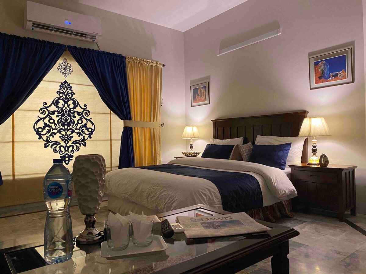 A serene bedroom is presented with a spacious bed dressed in crisp white linens and navy accents. Soft lighting is provided by two bedside lamps atop dark wood nightstands. Dark blue and gold curtains frame the window, enhancing the room's cozy and calming atmosphere.