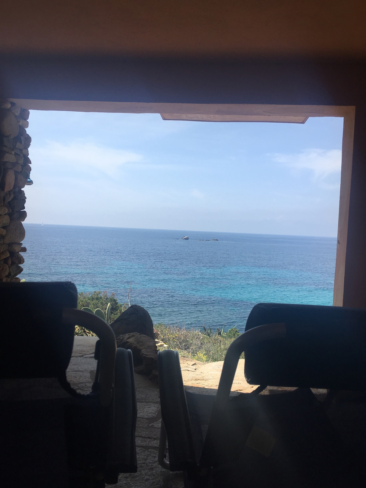 A serene view of the coastline and sea is framed by a stone wall. Two chairs are positioned in the foreground, offering a glimpse of the tranquil waters and clear blue sky beyond. The image evokes a sense of peaceful seclusion.