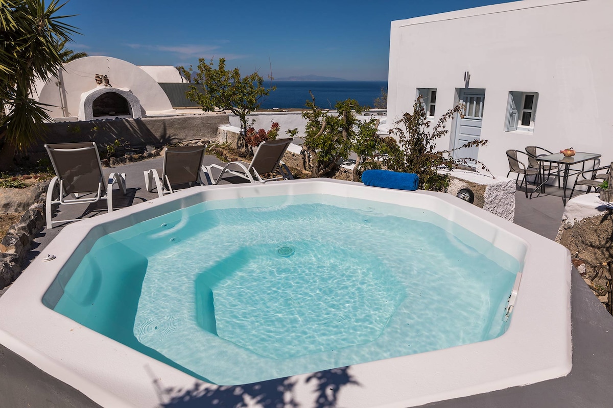 An inviting hot tub is presented in a private outdoor area, surrounded by lounge chairs. A table and additional seating are also visible, set against a backdrop of vibrant greenery and a clear view of the Aegean Sea.