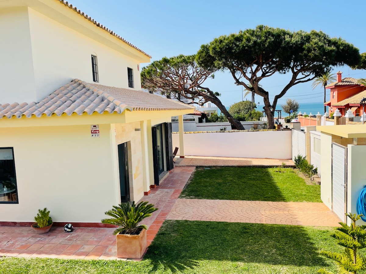A two-story house with a tiled roof is surrounded by greenery, including a well-maintained lawn. The sea is visible in the background, framed by large pine trees. A spacious driveway leads to the entrance, and a few decorative plants are positioned near the house.