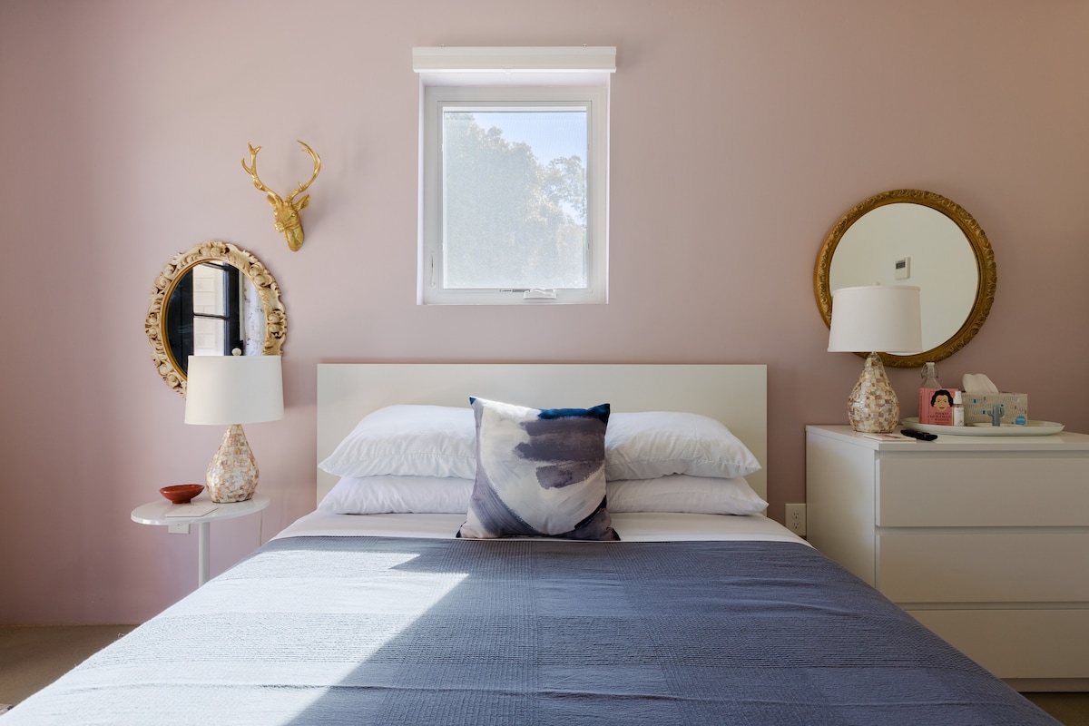 A comfortable bedroom features a spacious bed with soft linens. Light enters through a window, illuminating the soft pink walls. Decorative mirrors adorn each side of the bed, while bedside tables with lamps provide additional lighting. A subtle decorative accent pillow adds a touch of color.