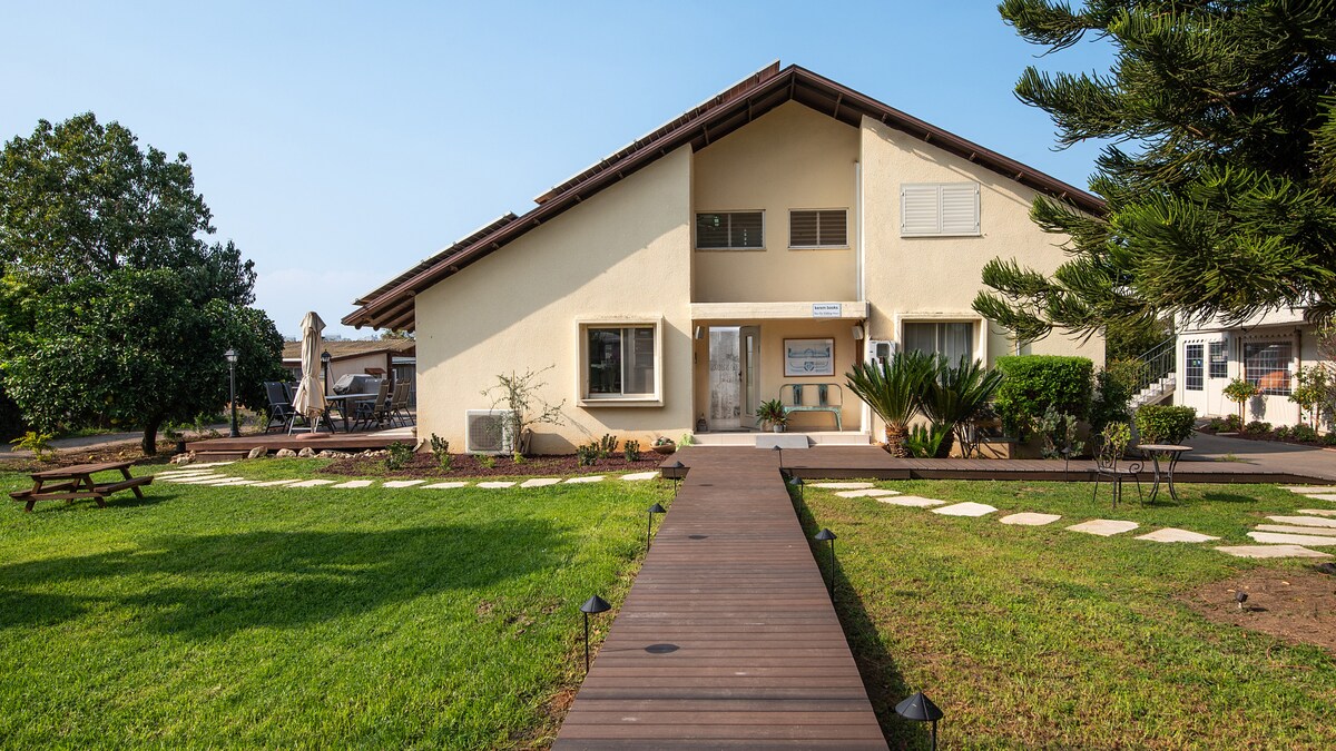 The front view of the house showcases a modern design with a light-colored exterior. A pathway leads to the entrance, bordered by well-maintained lawns and garden features. The surrounding area includes trees and outdoor seating, providing an inviting atmosphere.