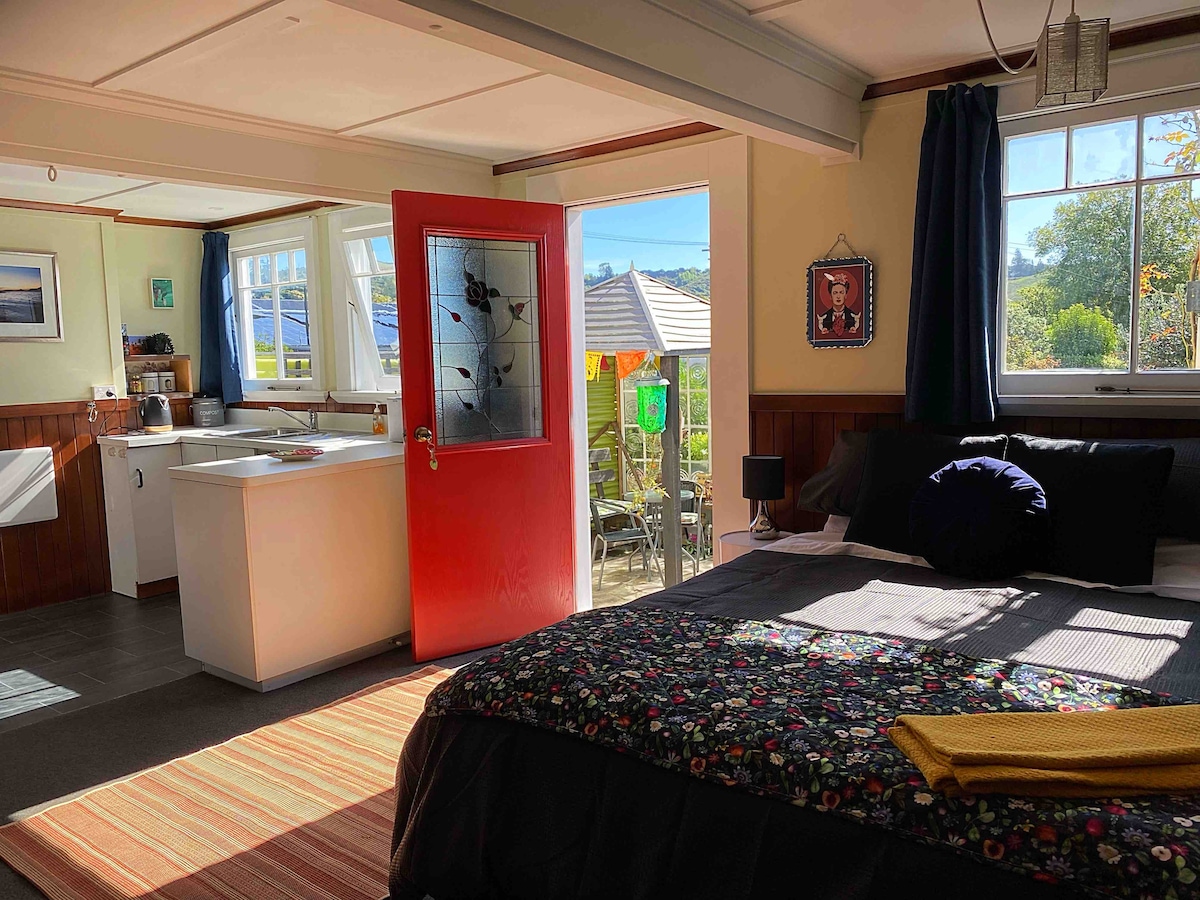 The studio space features a welcoming entrance with a vibrant red door, leading into a bright area that includes a spacious bed with a floral-patterned cover and a cozy black pillow. Natural light fills the room, highlighting an adjoining kitchenette and a view of the private garden.