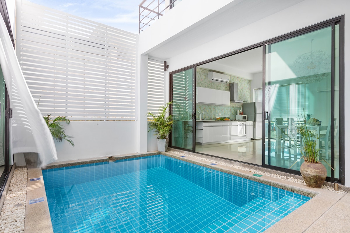 A private pool area is visible, surrounded by modern glass doors that open to a bright dining space. The pool features blue tiles, and tropical plants add greenery to the serene atmosphere. Natural light fills the area, enhancing the connection to the outdoor space.