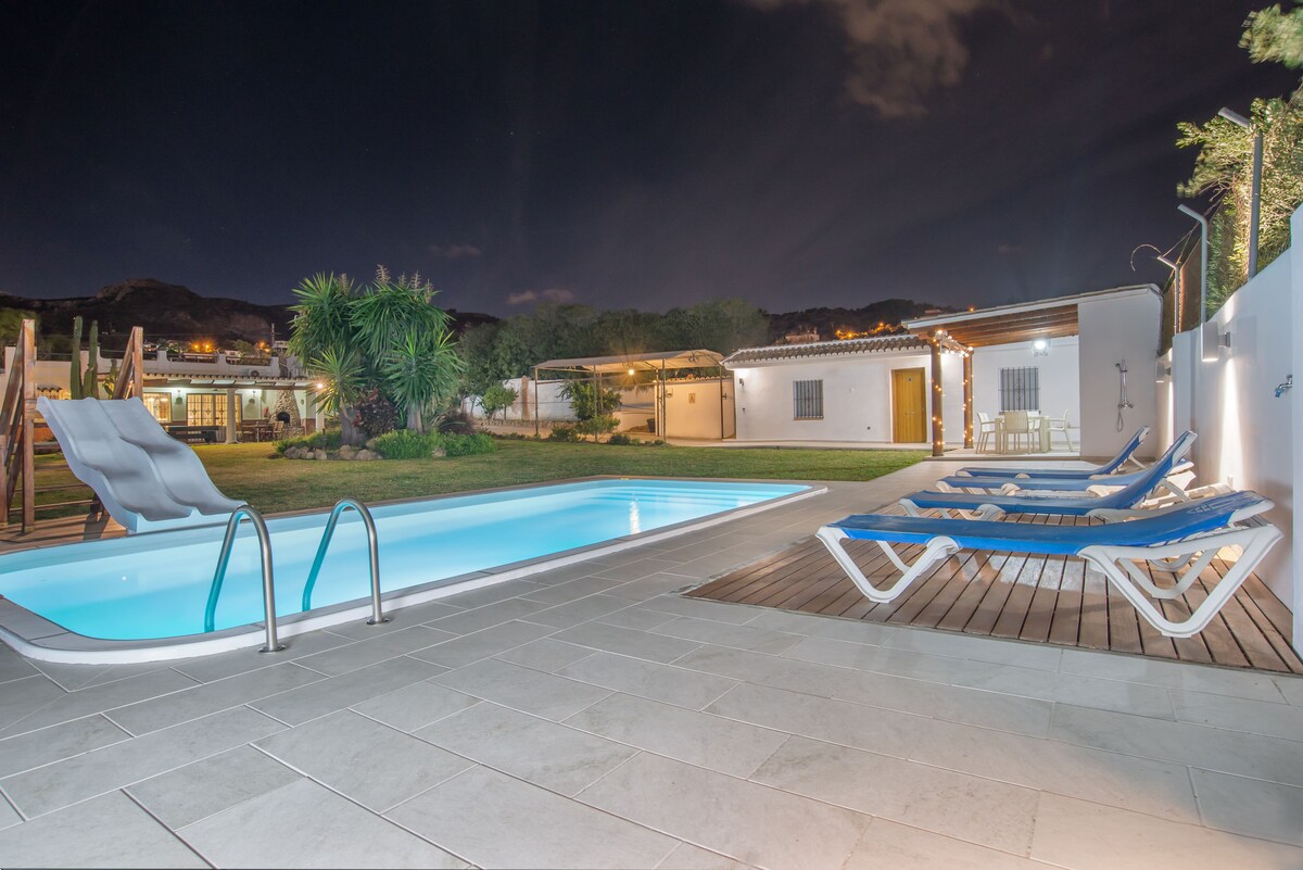 A private heated pool glows softly under nighttime lighting, surrounded by a spacious garden featuring palm trees and seating areas. Comfortable lounge chairs are positioned near the pool, while a slide adds a playful touch to the outdoor space.