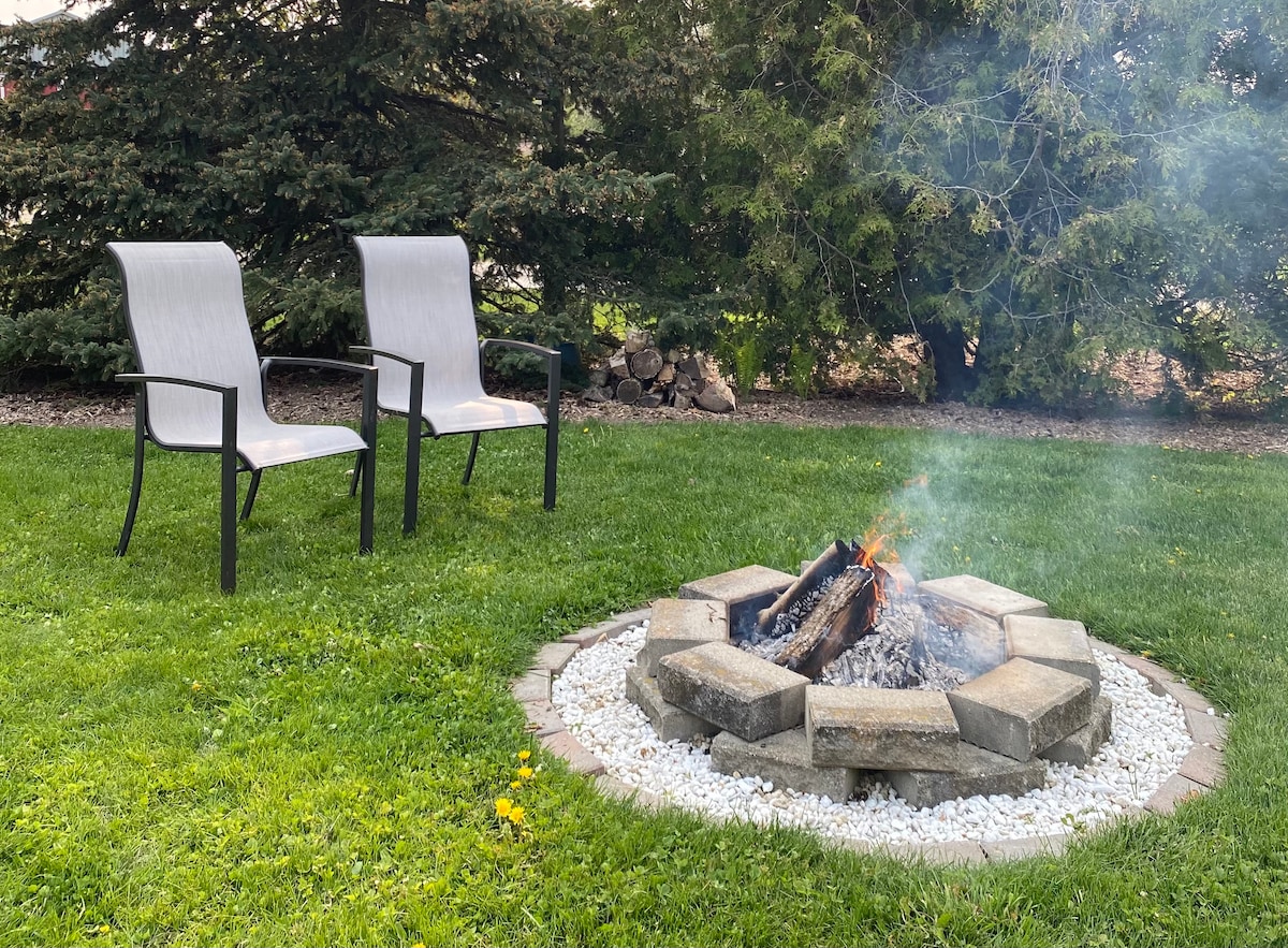 A stone firepit is surrounded by a circular arrangement of pavers and white gravel. Two gray chairs are positioned nearby. Lush green grass and shrubs create a natural backdrop, providing a serene outdoor setting.