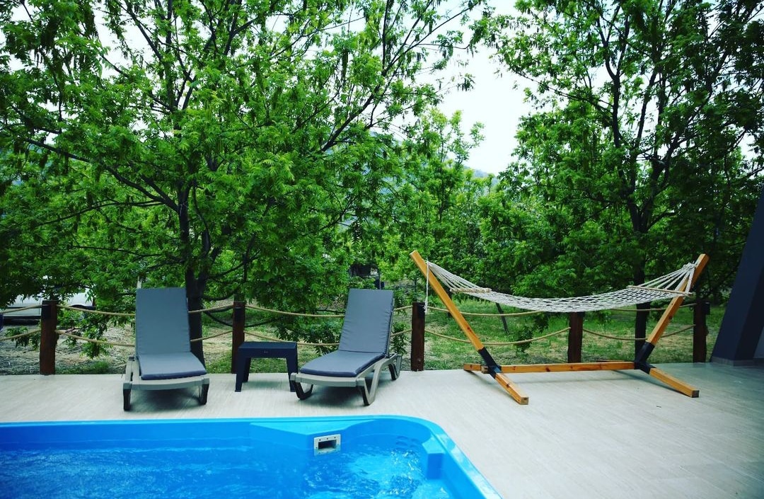 Poolside loungers are positioned next to a private pool, surrounded by lush green walnut trees. A cozy hammock is set nearby, inviting relaxation in the serene outdoor space. Natural light enhances the tranquil ambiance of this secluded garden area.