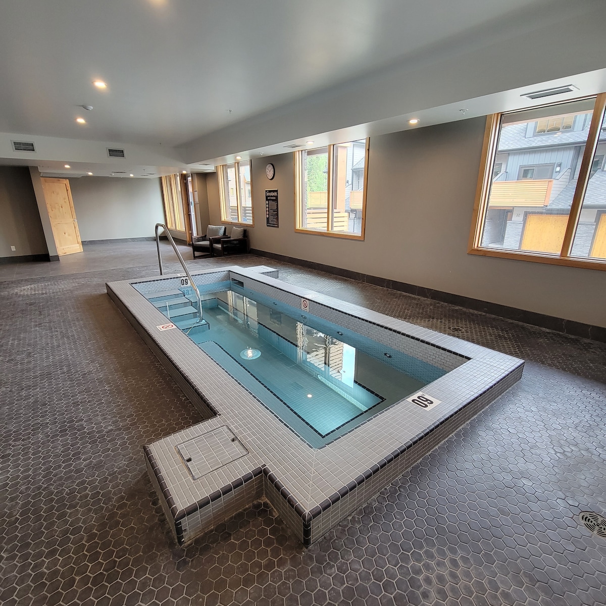 An indoor hot tub is situated within a spacious and well-lit area, surrounded by large windows allowing natural light. The tub features mosaic tile patterns and steps for easy access, with safety signage indicating depth. A clock is mounted on the wall for convenience.