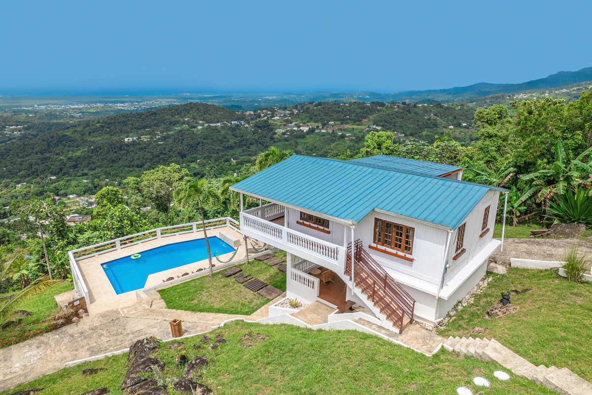 A scenic view of a two-level rustic house with a blue roof, surrounded by lush greenery and mountains. A swimming pool is visible in the front yard, adding to the serene landscape. The ocean can be seen in the distance under a clear blue sky.
