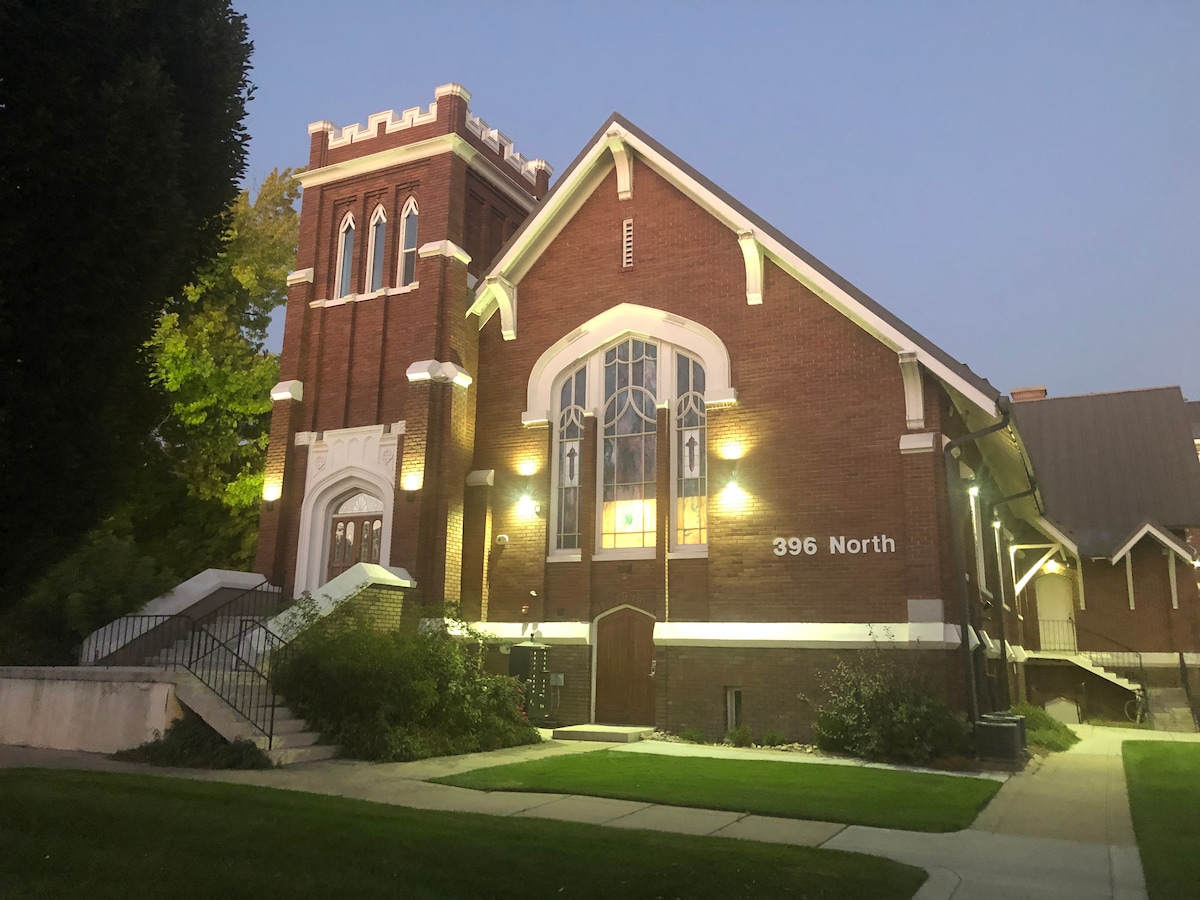 A historic church building is illuminated by soft lighting as dusk approaches. The red brick façade features large arched windows and a prominent entrance with white steps leading up to double doors. A manicured lawn surrounds the structure, enhancing its traditional charm.