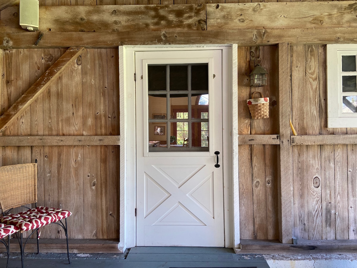 A white Dutch door is framed by rustic wooden walls, showcasing a combination of horizontal and vertical planks. A small lantern hangs nearby, and a woven basket adorns the wall beside the door. A simple bench with a patterned cushion sits on the side.