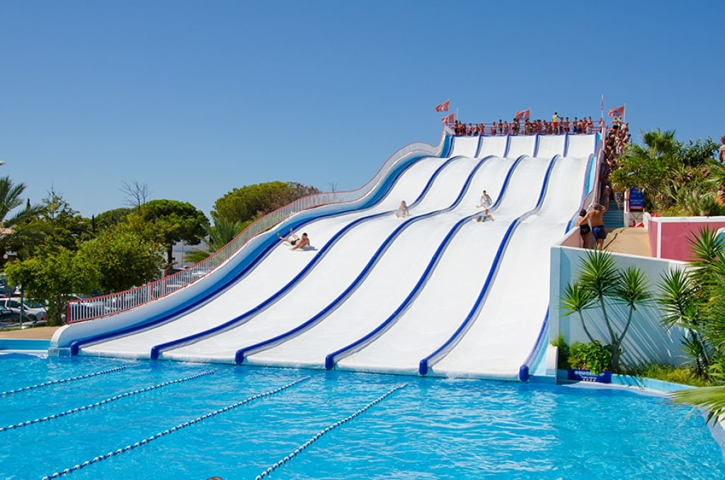 A large water slide structure is visible, featuring multiple lanes descending into a pool. Bright blue water reflects the clear sky. Surrounding the slide, palm trees and colorful landscaping create a lively atmosphere. People can be seen at the top, ready to slide down.