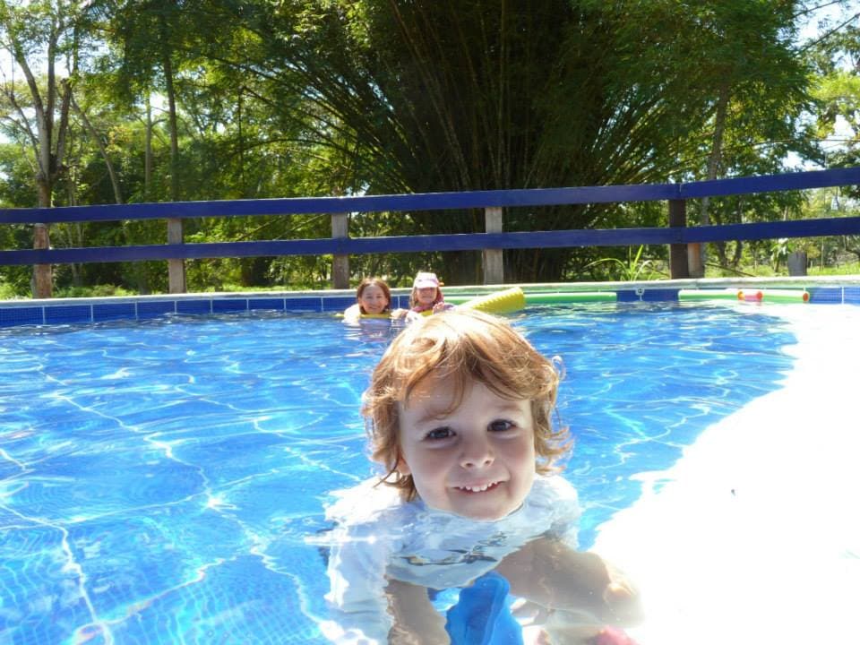 A child floats playfully in a clear blue pool surrounded by greenery. Sunlight reflects gently on the water's surface, creating a serene atmosphere. The pool's edge is lined with colorful pool floats, suggesting a fun and relaxing environment.