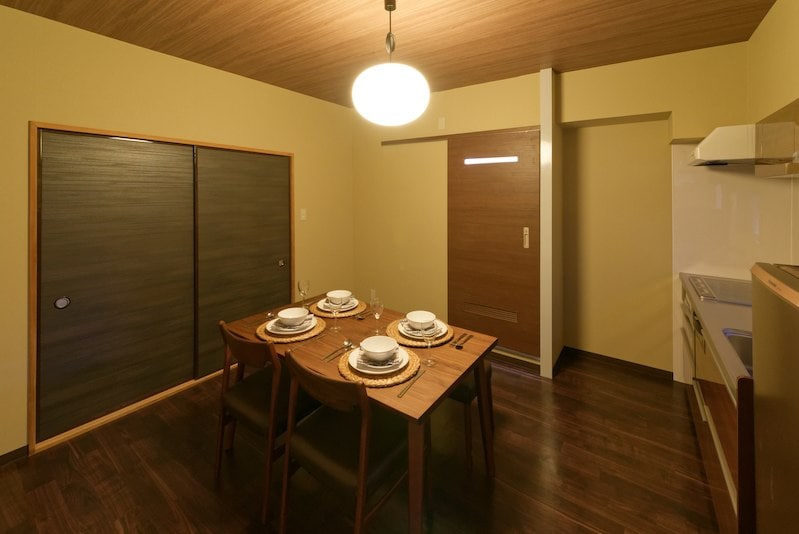 A spacious dining area is furnished with a wooden table set for four, including plates and utensils. Natural wood flooring complements the warm, neutral-toned walls. Nearby, a well-equipped kitchen is visible, featuring modern appliances and sleek cabinetry.