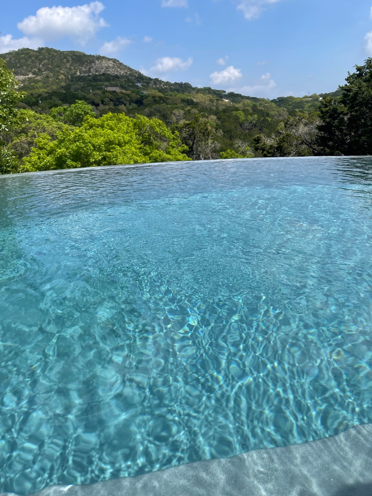 An infinity pool reflects the clear blue sky, surrounded by lush greenery and hills in the background. The water shimmers with sunlight, creating a tranquil and inviting atmosphere, perfect for relaxation.