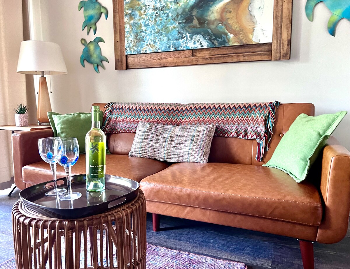 A cozy seating area features a brown leather couch adorned with patterned throw blankets and green accent pillows. A small round table holds two blue-tinted glasses and a green bottle, complemented by an art piece above the couch depicting ocean hues.