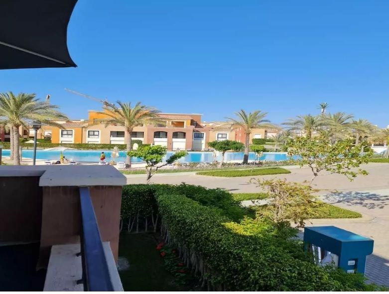 A view from a balcony showcases a clear blue sky and a well-maintained swimming pool surrounded by palm trees. The pool area features lounge chairs and is bordered by lush greenery, with buildings visible in the background that provide a residential atmosphere.