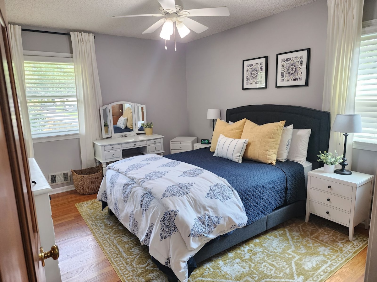 The bedroom features a spacious bed with a dark headboard, adorned with white and blue bedding. Two decorative pillows add a pop of color. Nightstands are placed on either side, and a dresser with a mirror occupies one corner. Natural light filters through window treatments.