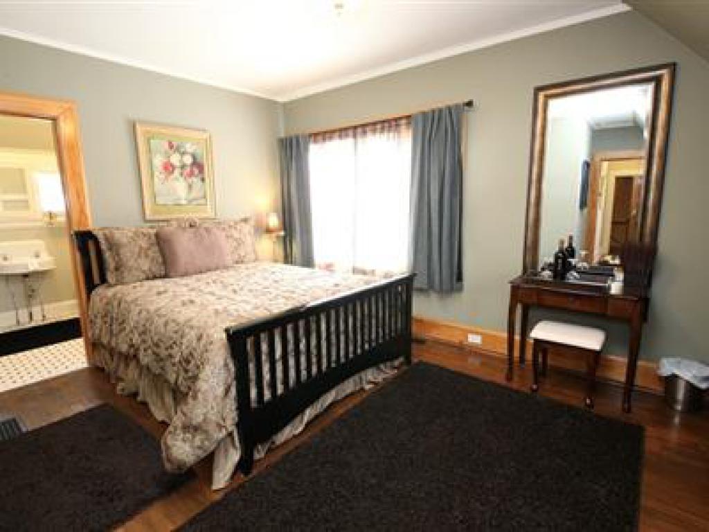A cozy bedroom features a Queen-sized bed with floral bedding, complemented by a plush area rug. A large mirror reflects the space, while natural light filters through draped curtains. An ensuite bathroom is visible through an open door in the background.
