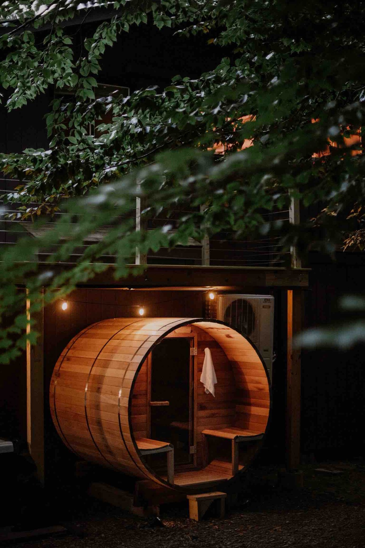A cedar barrel sauna is nestled in a shaded outdoor area, emphasizing natural wood tones. Soft lighting adorns the entrance, creating a cozy ambiance. Surrounding foliage adds a peaceful touch, while the structure's rounded shape offers a unique aesthetic complementing the tranquil setting.